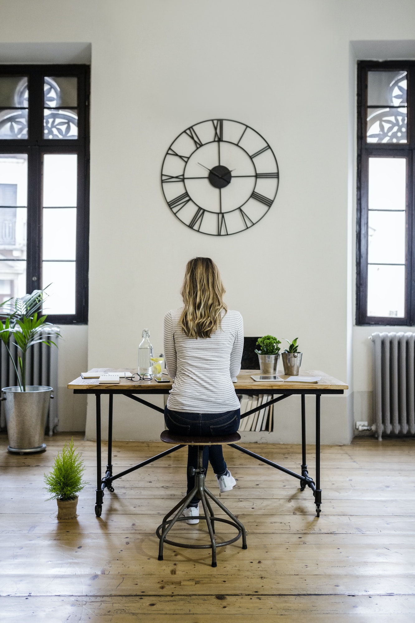 Rear view of woman sitting at desk at home under large wall clock