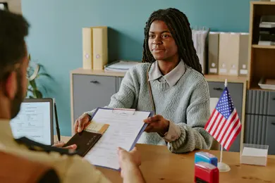 African American woman holding clipboard and pen during visa interview. Office setting with documents and American flag on desk and bookshelf in background